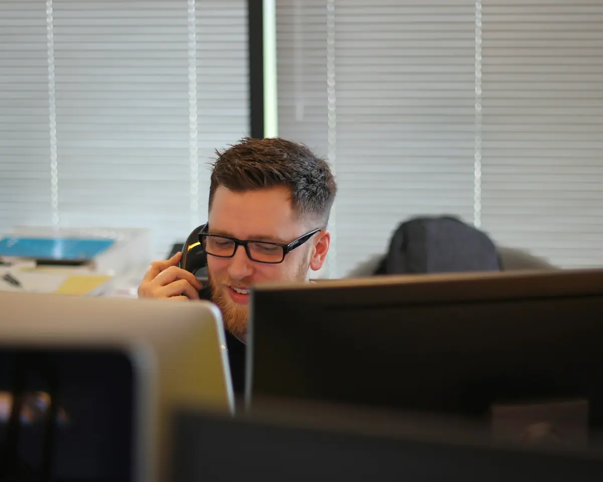 Vendedor fala ao telefone na sua mesa de trabalho, entre monitores do escritório